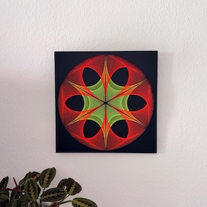 Puede incluir: Una pieza de arte de hilo geométrico con un diseño circular de hilos rojos y verdes sobre un cuadrado negro. La obra de arte está montada en una pared blanca, creando una estética moderna. Dimensiones: aproximadamente 30 cm x 30 cm.