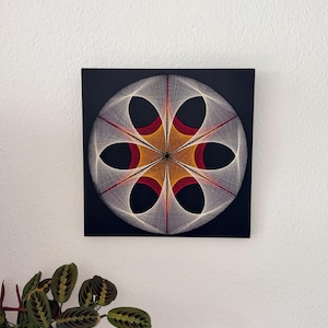 Può includere: Arte geometrica con filo su sfondo nero quadrato. Il design presenta un motivo simmetrico di fili bianchi, rossi e dorati, creando una forma a fiore. L'opera d'arte è montata su una parete, con una pianta in vaso in primo piano.