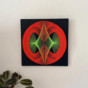 Op de afbeelding: Een geometrisch draadkunstwerk op een zwart vierkant canvas. Het ontwerp heeft een centrale ruitvorm in groen, omgeven door twee rode cirkels. Het kunstwerk is aan een witte muur bevestigd, met een groene plant in de linkerbenedenhoek.
