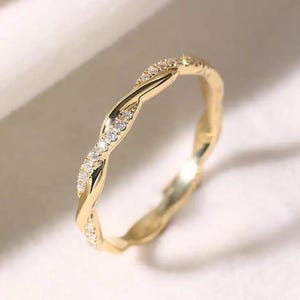 May include: A gold-colored ring with a twisted design. The ring is adorned with small, clear gemstones set along the twist. The ring is displayed on a white surface, highlighting its intricate details and elegant appearance.