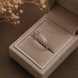 Puede incluir: Un anillo de tono plateado con tres engastes en forma de marquesa, cada uno con una peque&ntilde;a piedra transparente y brillante. El anillo est&aacute; colocado en &aacute;ngulo dentro de un joyero rectangular de color beige forrado de terciopelo.