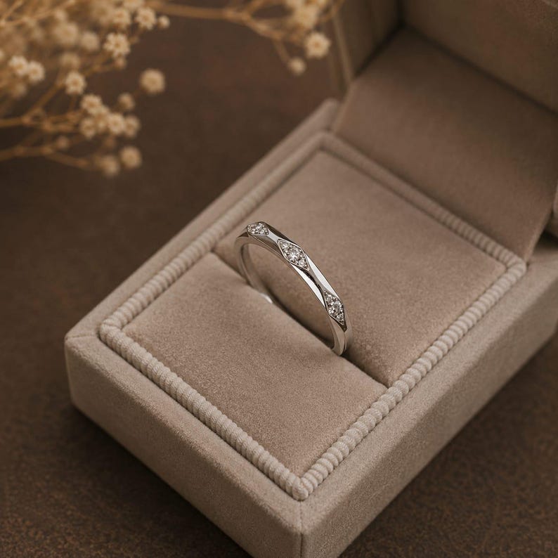 Puede incluir: Un anillo de tono plateado con tres engastes en forma de marquesa, cada uno con una peque&ntilde;a piedra transparente y brillante. El anillo est&aacute; colocado en &aacute;ngulo dentro de un joyero rectangular de color beige forrado de terciopelo.