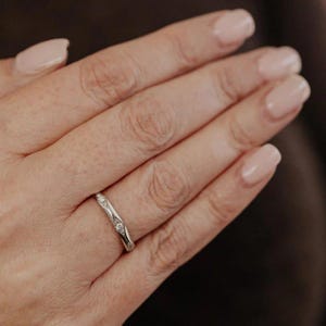 Puede incluir: Un anillo de banda de metal plateado que presenta una serie de peque&ntilde;as piedras redondas y transparentes engastadas en monturas individuales en forma de diamante a lo largo de la superficie exterior. El anillo tiene un acabado pulido y fino.