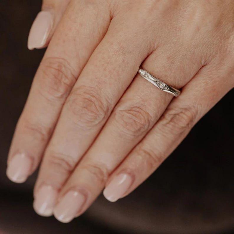 Puede incluir: Un anillo de banda en tono plateado con un perfil ligeramente curvado y ondulado. La superficie de la banda met&aacute;lica est&aacute; acentuada con tres peque&ntilde;as piedras redondas y transparentes engastadas a intervalos a lo largo de la circunferencia.