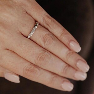 Puede incluir: Un anillo de metal en tono plateado con una banda delgada y un contorno ligeramente ondulado. La banda est&aacute; engastada con tres peque&ntilde;as piedras redondas y transparentes dispuestas en fila a lo largo del centro, con acabado pulido.