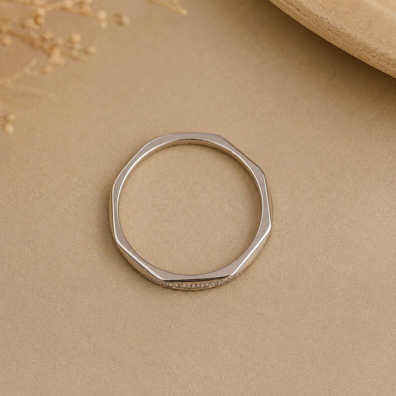 Puede incluir: Un anillo de tono plateado con un dise&ntilde;o de banda geom&eacute;trica facetada. La superficie exterior del anillo presenta una fila fina y continua de peque&ntilde;as piedras claras y brillantes incrustadas a lo largo de una secci&oacute;n de la banda de metal.