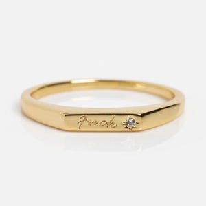 May include: A gold-toned ring featuring a flat, rectangular bar section on the front. The bar is engraved with the cursive text "fuck" and is accented with a small, clear, star-shaped inset stone positioned to the right of the lettering.