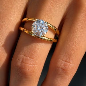 Op de afbeelding: Een gouden ring met een enkele ronde, briljant geslepen heldere edelsteen, geplaatst tussen twee parallelle, gepolijste gouden banden. Het ontwerp creëert een open ruimte rond de middelste steen, wat de facetten van de edelsteen benadrukt.