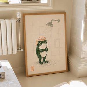 May include: A framed print featuring a green frog with a light orange shower cap standing in a tiled shower under a showerhead. The frog has its arms crossed and a grumpy expression. There is a small red square stamp with Japanese text.