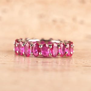 Pode incluir: Um anel de eternidade em tom prateado com uma fila contínua de pedras preciosas ovais em rosa vibrante. Cada pedra é fixada por pequenas garras de metal polido, criando um design circular e uniforme ao redor de todo o anel.