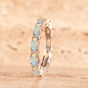 Puede incluir: Un anillo de eternidad en tono plateado que presenta una serie de ópalos blancos iridiscentes de forma ovalada dispuestos en fila vertical. Pequeñas piedras redondas y transparentes se sitúan entre cada ópalo, creando una banda continua.