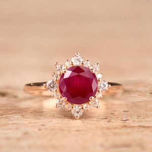 Könnte beinhalten: Ein Ring aus Roségold mit einem großen, runden, tiefroten Rubin als Mittelstein, der von einem Kranz aus kleinen, klaren, rund geschliffenen weißen Diamanten umgeben ist. Die Fassung ist kunstvoll, mit sternförmig angeordneten Diamanten.