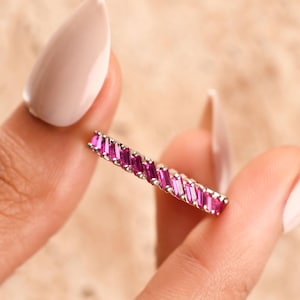 Könnte beinhalten: Ein dünner silberfarbener Ring mit einer durchgehenden Reihe von leuchtend magentafarbenen Baguette-Steinen, die horizontal gefasst sind. Die Steine sind in einem sich wiederholenden Muster entlang des gesamten Metallrings angeordnet.