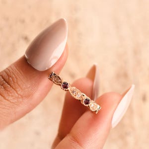 May include: A rose gold band ring featuring an alternating pattern of small round dark red garnet gemstones and clear white diamonds set within a delicate milgrain textured metal frame that creates a repeating marquise-shaped design.