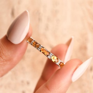 Op de afbeelding: Een dunne ring met een reeks afwisselende ovaal geslepen gele citrien edelstenen en kleine heldere witte steentjes in een metalen zetting. Het ontwerp toont een herhalend patroon van oranje-gele stenen geaccentueerd door kleine heldere steentjes.
