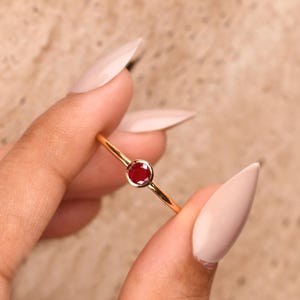 Puede incluir: Un anillo fino de oro con una sola piedra preciosa de color rojo intenso, redonda y facetada, engastada en un bisel liso. La banda es delgada y pulida, mostrando un diseño sencillo que resalta el color vibrante de la piedra central.