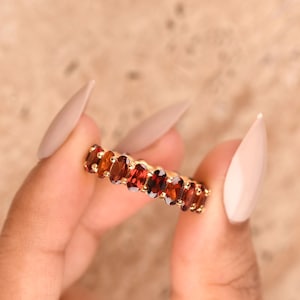 May include: A gold eternity band featuring a continuous row of oval-cut, deep red garnet gemstones set in a prong setting. The ring displays a vibrant gradient of dark red to orange-red hues across the faceted stones, creating a uniform circular design.