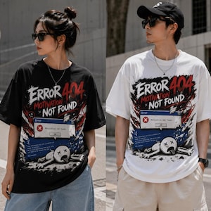 Op de afbeelding: Zwarte en witte T-shirts met de tekst "ERROR 404 MOTIVATION NOT FOUND" in een verweerde lettertype. Het ontwerp bevat een computerfoutmelding en een cartoonfiguur. De shirts zijn korte mouwen.