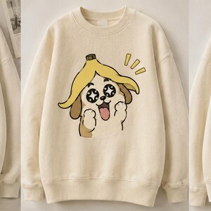 Puede incluir: Sudadera de color crema con un perro de dibujos animados que lleva un sombrero de cáscara de plátano. La expresión del perro varía en las tres sudaderas, una sonriendo, otra emocionada y otra sorprendida. Las sudaderas se muestran en perchas de madera.