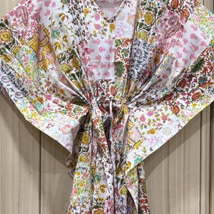 May include: A white cotton kaftan featuring a vibrant multi-coloured floral and paisley block print pattern. The garment has a V-neckline, wide kimono-style sleeves, and a drawstring waist tie that gathers the fabric to create a draped silhouette.
