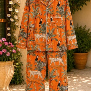 May include: A long-sleeved button-down shirt and matching trousers set in a vibrant orange fabric. The garment features a repeating pattern of cheetahs, toucans, and palm trees, accented with white piping along the collar, cuffs, and pockets.