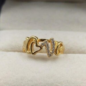 Puede incluir: Un anillo de tono dorado que forma la palabra LOVE en una fuente cursiva estilizada. La letra O tiene forma de corazón, y las letras L, V y E están adornadas con pequeñas piedras claras y brillantes incrustadas en la superficie del metal.