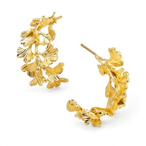 May include: A pair of gold hoop earrings featuring a cluster of textured ginkgo leaf motifs arranged along the curved band. Each earring has a post back closure and a polished metallic finish that reflects light across the detailed fan-shaped leaves.