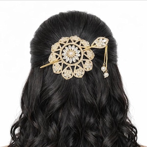 Könnte beinhalten: Ein goldfarbenes Haaraccessoire mit einem kreisförmigen, blumenartigen Design, das mit klaren, marquise-geschliffenen Kristallen und einer zentralen Perle besetzt ist. Eine dünne goldene Nadel verläuft durch die Mitte, mit zwei zarten Ketten.