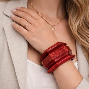 Puede incluir: Un conjunto de tres brazaletes gruesos de resina de color rojo intenso con un diseño geométrico facetado en la muñeca, junto con un collar de cadena fina dorada con un pequeño colgante circular y un anillo de banda dorada.