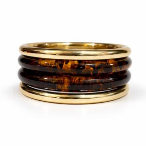 Puede incluir: Un anillo de varias bandas con dos aros de metal en tono dorado pulido en la parte superior e inferior, que encierran dos bandas centrales de resina con patrón de carey en tonos marrón oscuro y ámbar, creando un aspecto apilado.