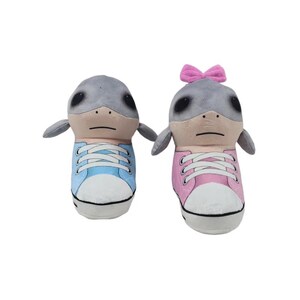 K&ouml;nnte beinhalten: Zwei Pl&uuml;schtiere in Form von Robben, die jeweils einen Turnschuh tragen. Einer hat einen blauen Turnschuh, der andere einen rosa Turnschuh mit einer rosa Schleife. Die Robben haben graue K&ouml;pfe, kleine schwarze Augen und kleine graue Flossen.