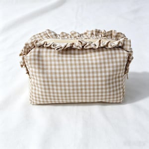 May include: A rectangular zipped pouch featuring a beige and white gingham check pattern. The top edge of the pouch is decorated with a gathered fabric ruffle in the same matching gingham print, extending across the length of the zip closure.
