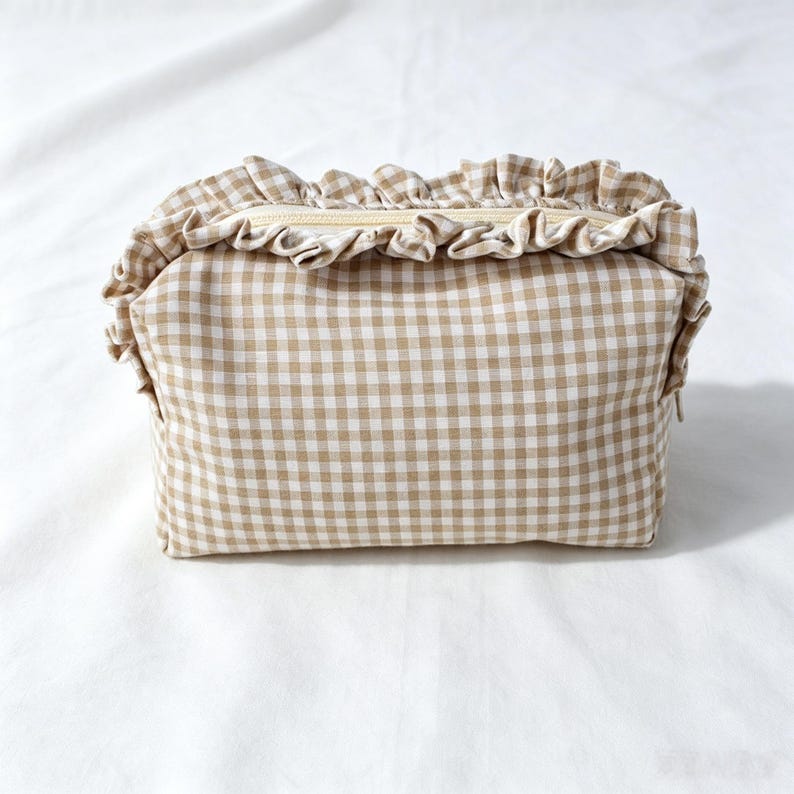 May include: A rectangular zipped pouch featuring a beige and white gingham check pattern. The top edge of the pouch is decorated with a gathered fabric ruffle in the same matching gingham print, extending across the length of the zip closure.