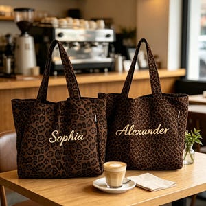 May include: Two matching tote bags featuring a dark brown and black leopard print pattern. Each bag has two shoulder straps and is personalised with embroidered text in a light tan script font, reading Sophia on one bag and Alexander on the other.