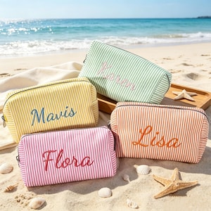 Puede incluir: Cuatro estuches rectangulares con cremallera y rayas verticales en verde claro, amarillo, rosa y naranja. Cada estuche presenta nombres bordados en letra cursiva: Karen, Mavis, Flora y Lisa. Los estuches están sobre la arena de la playa.