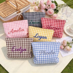May include: Six rectangular zippered pouches in various gingham patterns, each embroidered with a unique name: Amelia in pink, Lucas in black, Harper in red, Logan in yellow, Evelyn in brown, and Phoebe in blue. The pouches are arranged together.