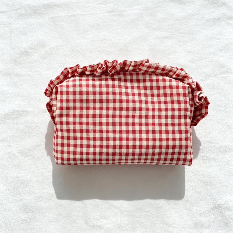 May include: A rectangular zipped pouch featuring a red and white gingham check pattern. The pouch is accented with a gathered fabric ruffle trim along the top edge, creating a soft, textured border around the structured, boxy main compartment.