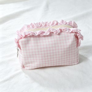 May include: A rectangular zipped pouch featuring a light pink and white gingham check pattern. The top edge of the pouch is decorated with a gathered fabric ruffle in the same light pink gingham material, and it includes a silver metal zip pull.