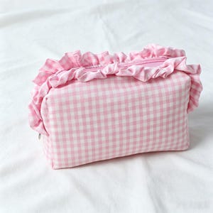 May include: A rectangular zipped pouch featuring a light pink and white gingham check pattern. The top edge of the pouch is decorated with a gathered fabric ruffle made from the same pink and white checkered material, surrounding the zip.