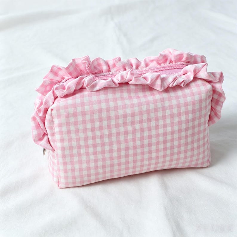 May include: A rectangular zipped pouch featuring a light pink and white gingham check pattern. The top edge of the pouch is decorated with a gathered fabric ruffle made from the same pink and white checkered material, surrounding the zip.