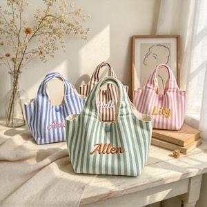 May include: A collection of four small canvas tote bags featuring vertical stripes in blue, brown, green, and pink. Each bag has a single button closure and is embroidered with a name: Arie, Hall, Allen, and Lily in contrasting thread colours.