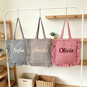 May include: Three square tote bags with ruffled edges and a gingham pattern in black, brown, and red. Each bag features a personalized name embroidered in the center: Sofia on the black bag, Amelia on the brown bag, and Olivia on the red bag.