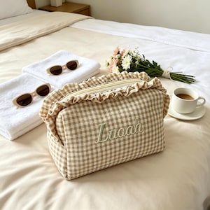 May include: A beige and white gingham patterned cosmetic bag with a ruffled trim along the top zip. The name "Lucaah" is embroidered in light green thread on the front of the bag. The bag features a rectangular shape and a soft fabric texture.