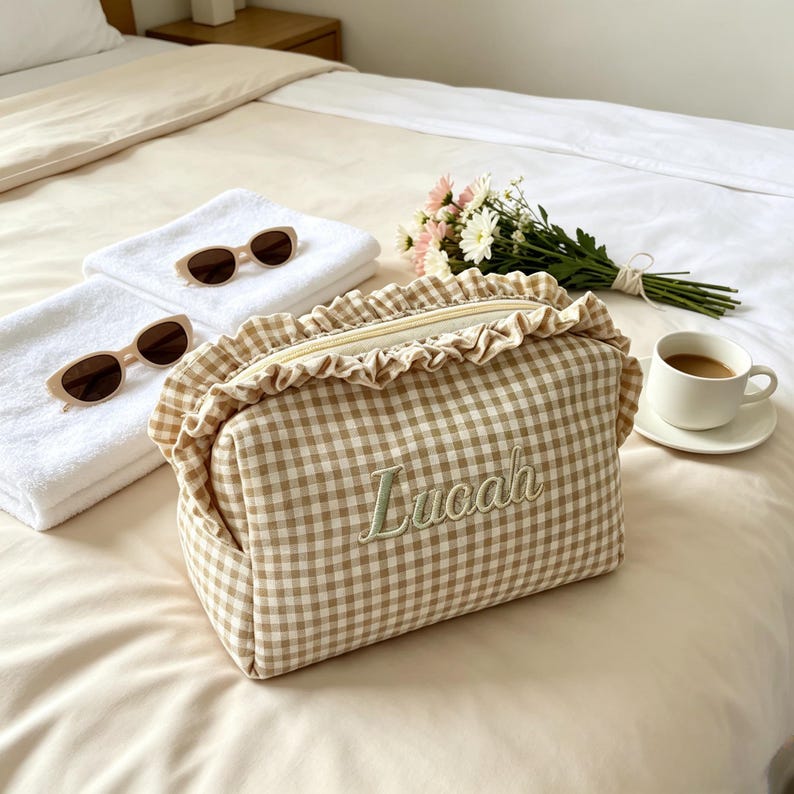 May include: A beige and white gingham patterned cosmetic bag with a ruffled trim along the top zip. The name "Lucaah" is embroidered in light green thread on the front of the bag. The bag features a rectangular shape and a soft fabric texture.