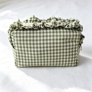 May include: A rectangular zipped pouch featuring a green and white gingham check pattern. The top edge of the pouch is adorned with a gathered fabric ruffle in the same green and white checkered material, extending across the top closure.