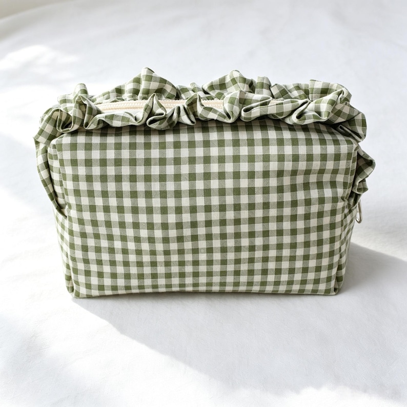 May include: A rectangular zipped pouch featuring a green and white gingham check pattern. The top edge of the pouch is adorned with a gathered fabric ruffle in the same green and white checkered material, extending across the top closure.