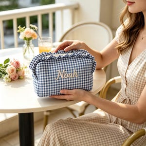 May include: A rectangular fabric pouch featuring a blue and white gingham check pattern with a ruffled top edge. The name Noah is embroidered in white script lettering on the front centre of the pouch, which has a zip closure.