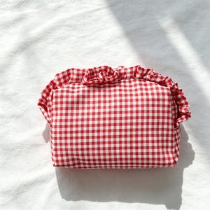 May include: A rectangular pouch featuring a red and white gingham check pattern. The pouch is accented with a ruffled fabric trim that runs along the top edge, creating a gathered, decorative border across the front of the accessory.