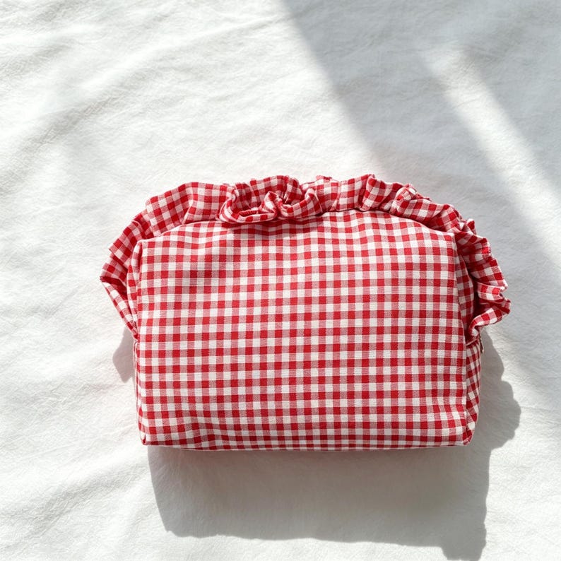 May include: A rectangular pouch featuring a red and white gingham check pattern. The pouch is accented with a ruffled fabric trim that runs along the top edge, creating a gathered, decorative border across the front of the accessory.