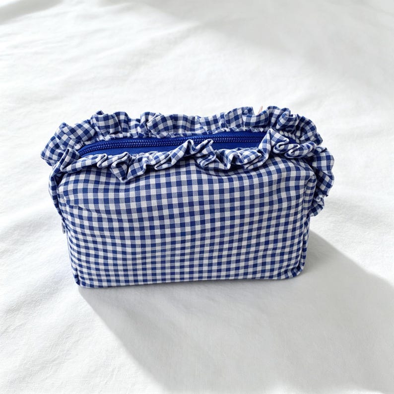 May include: A rectangular zipped pouch featuring a blue and white gingham check pattern. The top opening is accented with a gathered fabric ruffle in the same blue and white checkered material, and the zip closure is a solid royal blue colour.